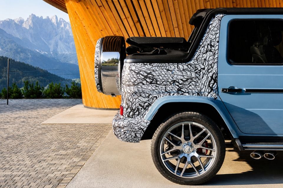 Rear three-quarter view of a blue 2027 Mercedes-AMG G63 Cabriolet with camouflage wrap on the rear, parked near a timber structure with mountains in the background