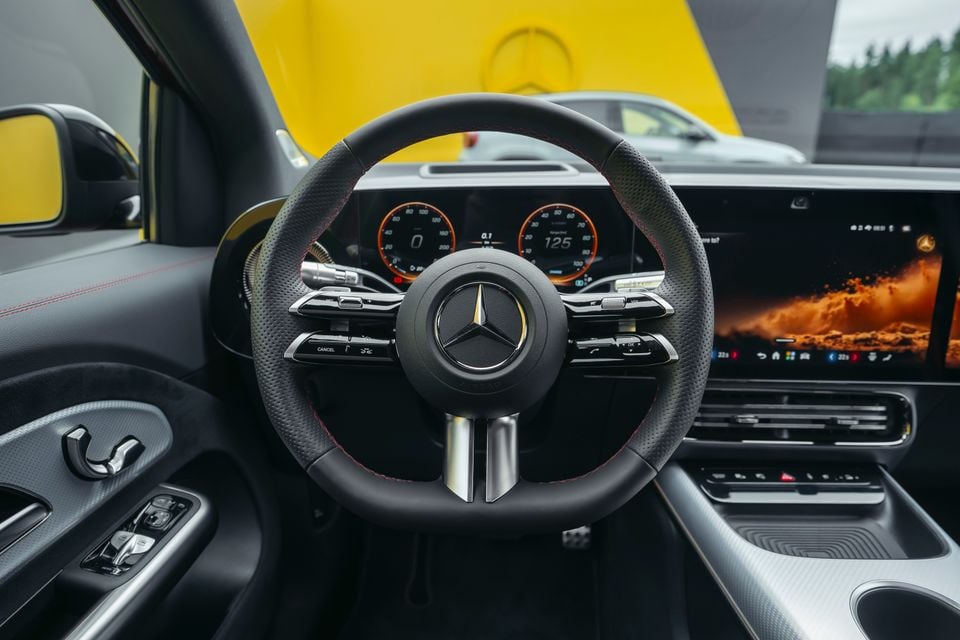 Mercedes-Benz GLA interior showing the leather-trimmed steering wheel with AMG-style red stitching, digital instrument cluster with orange dials, and a large infotainment touchscreen