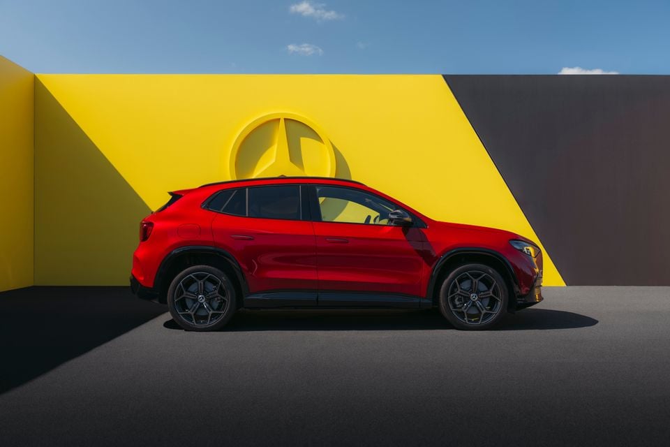A red Mercedes-Benz GLA SUV seen from the side, parked in front of a yellow and dark geometric backdrop featuring a large Mercedes-Benz star logo.
