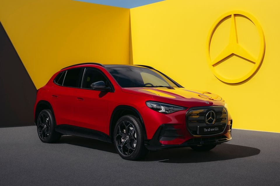 A red 2027 Mercedes-Benz GLA SUV seen from the front three-quarter angle, posed in front of a yellow backdrop featuring the Mercedes-Benz star logo.