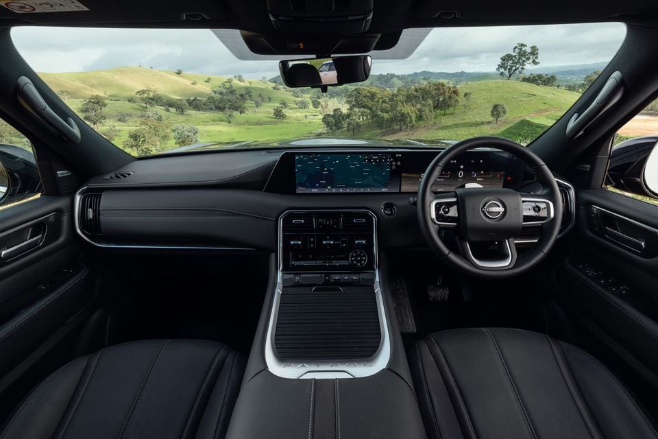 Interior view of the Nissan Patrol Y63 showing the dashboard, large infotainment touchscreen, digital instrument cluster, steering wheel, and centre console, with green Australian hills visible through the windscreen