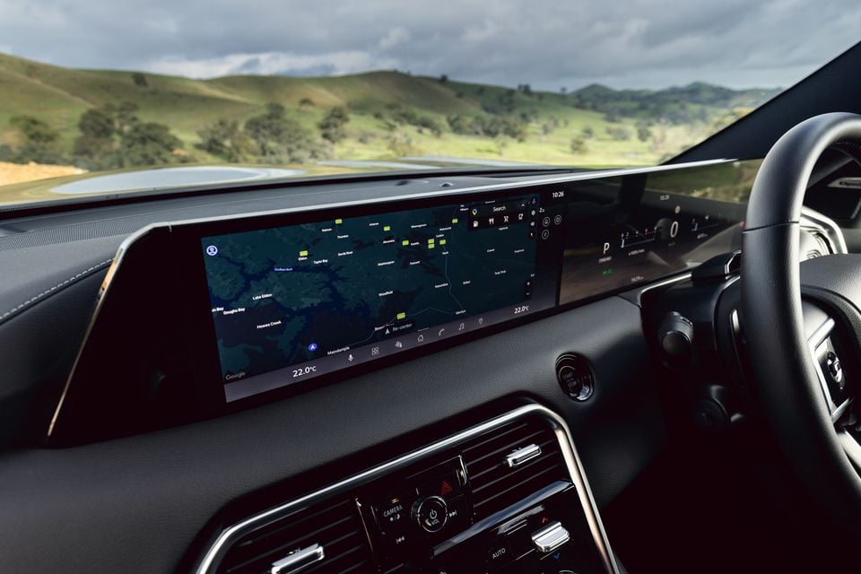 Interior of a 2027 Nissan Patrol Y63 showing the wide touchscreen infotainment display with Google Maps navigation, dashboard controls, and steering wheel, with green hills visible through the windscreen.