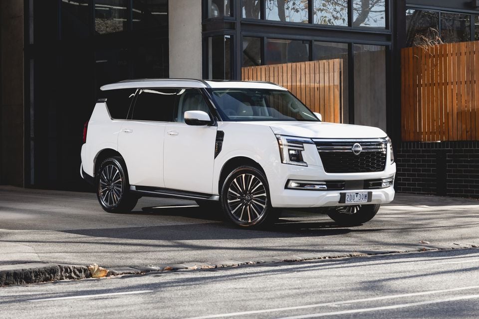 A white Nissan Patrol large SUV seen from the front-side angle, parked on a street beside a modern building