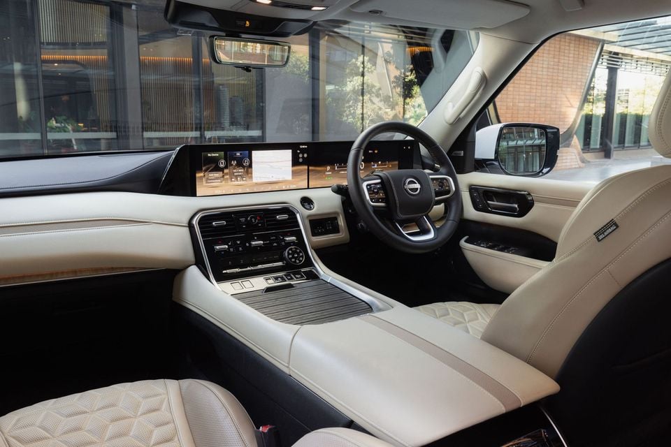 Nissan Patrol interior showing the widescreen infotainment display, Nissan-badged steering wheel, cream leather seats and centre console, viewed from the rear passenger side