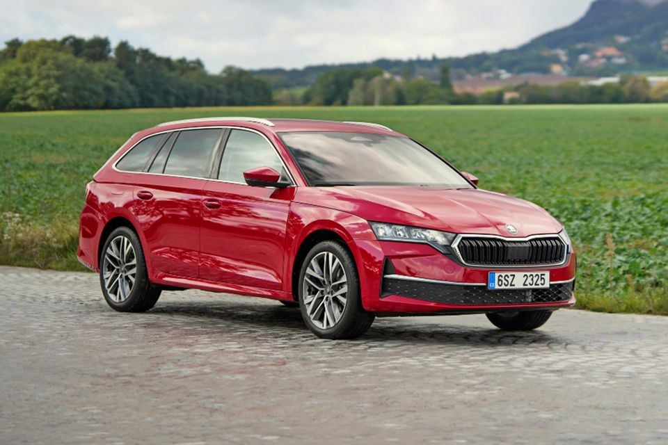 A red Skoda Octavia wagon parked on a paved road beside a green field, seen from the front quarter.