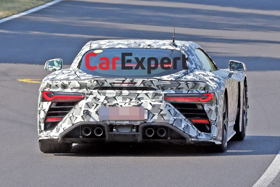 A camouflaged supercar prototype, believed to be the Toyota GR GT, seen from the rear on a race track, showing quad exhausts and wide body styling