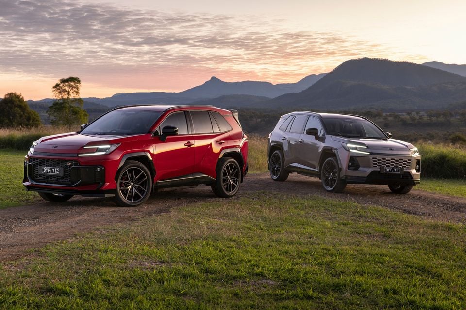 Two 2027 Toyota RAV4 GR Sport SUVs, one red and one silver, parked on grass at sunset with mountains in the background
