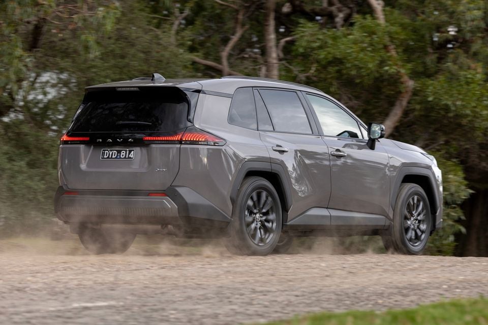 A grey Toyota RAV4 SUV kicks up dust as it drives along a gravel road, seen from the rear quarter.