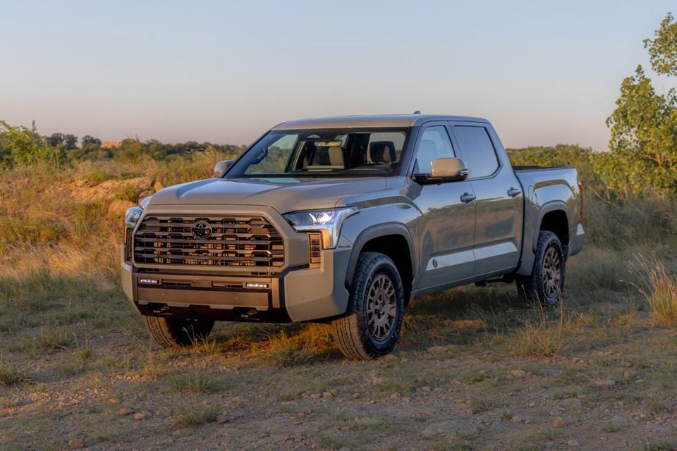 A Toyota Tundra Trailhunter crew-cab ute in a sandy tan colour, seen from the front-side angle on a dirt trail surrounded by dry grass and scrub.