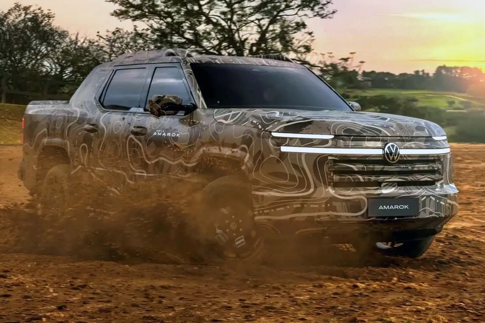 A Volkswagen Amarok ute prototype in camouflage wrap driving on a dirt surface, seen from the front-side at sunset