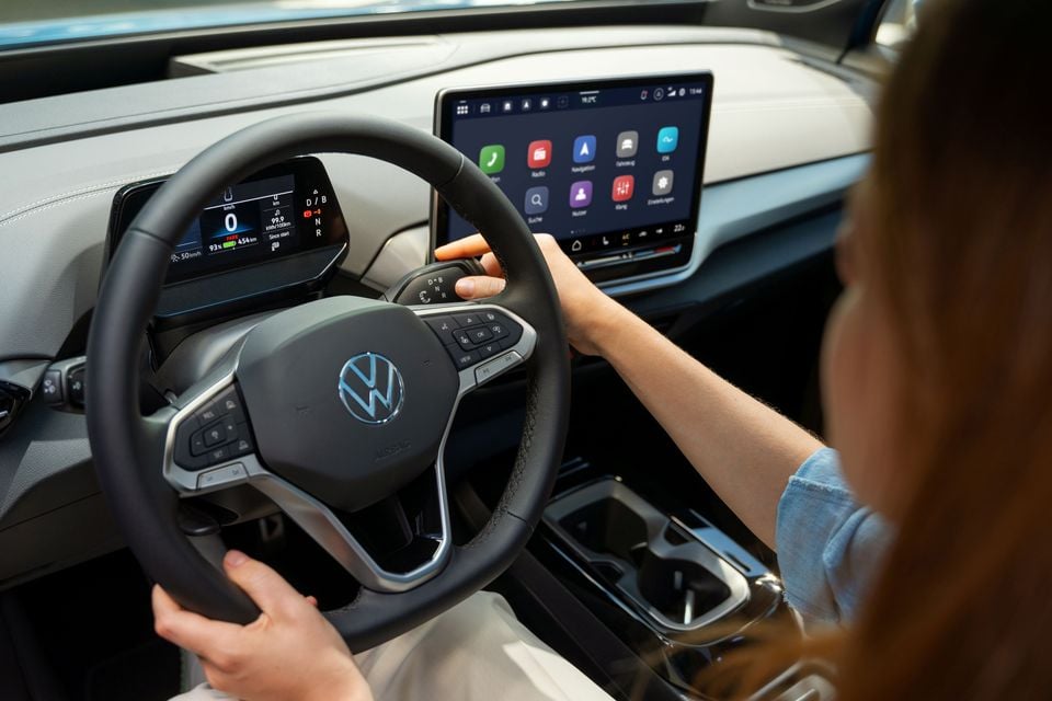 Driver's view of the 2027 Volkswagen ID.4 dashboard, with a hand on the steering wheel and gear selector beside the digital driver display and central touchscreen