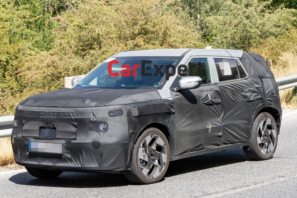 A heavily camouflaged Hyundai Kona SUV prototype seen from the front-side angle on a road, with foliage in the background
