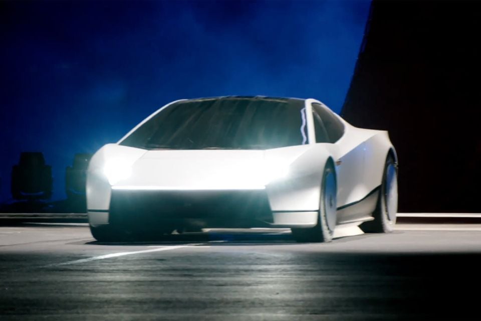 A white 2028 Tesla Roadster with its headlights on, seen from the front three-quarter angle on a dimly lit stage