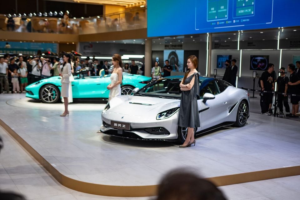 A white Denza Z sports car on display at a motor show, with a teal Denza Z convertible visible in the background and show models standing nearby