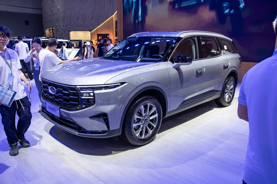 A silver Ford Edge L SUV on display at a motor show, seen from the front-side angle, with attendees browsing nearby.