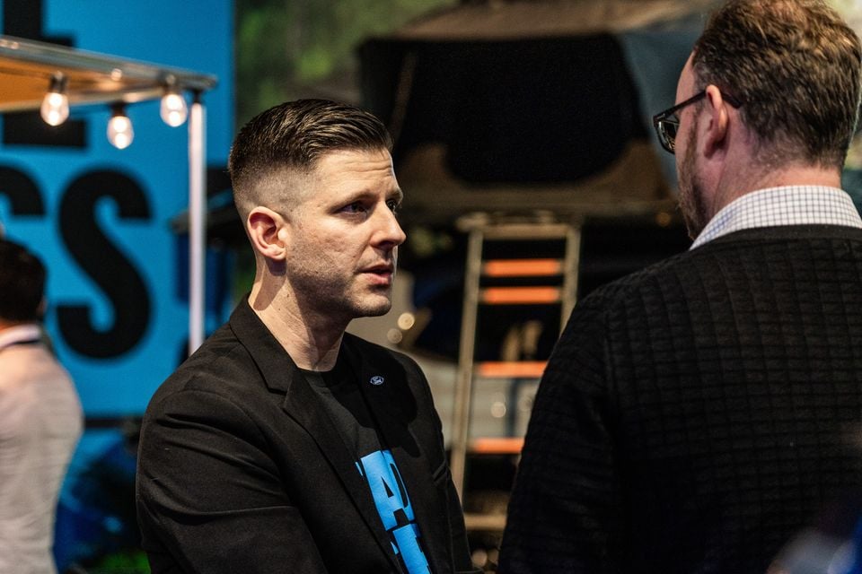 Two men in conversation at what appears to be an automotive industry event or motor show, with a blue branded display and roof tent visible in the background.
