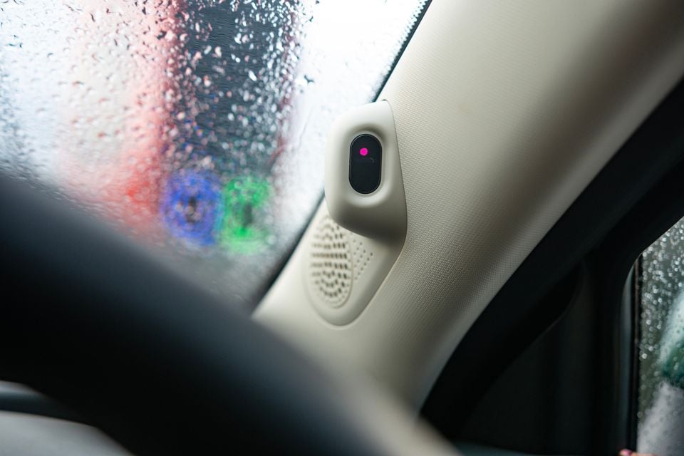 Interior view of a car's A-pillar showing a driver monitoring camera sensor with a pink indicator light, beside a speaker grille, with a rain-covered windscreen visible
