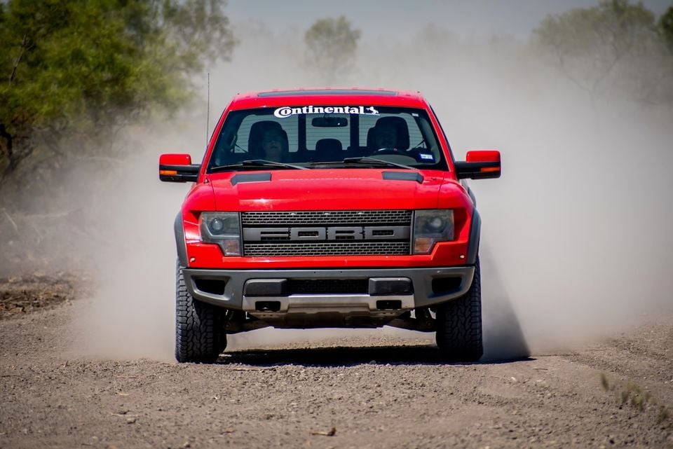 A red Ford F-150 SVT Raptor ute driving head-on along a dusty dirt track, kicking up a cloud of dust behind it