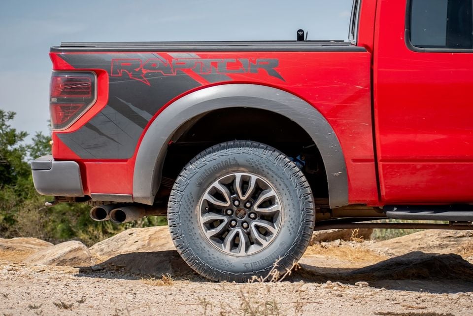 Close-up of the rear quarter panel and wheel of a red Ford F-150 SVT Raptor ute, showing the Raptor graphic on the tub and a Continental all-terrain tyre on rocky terrain