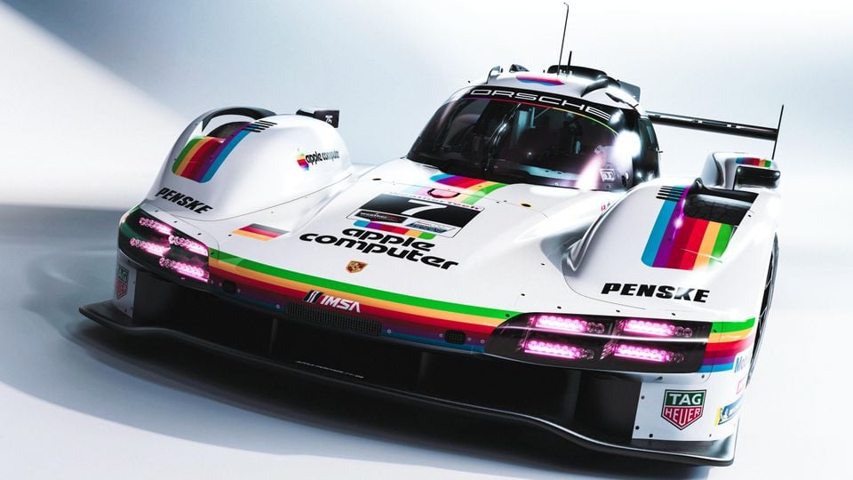A Porsche 963 LMDh racing car in a white livery featuring rainbow Apple Computer and Penske branding, seen from the front at a low angle in a studio setting.