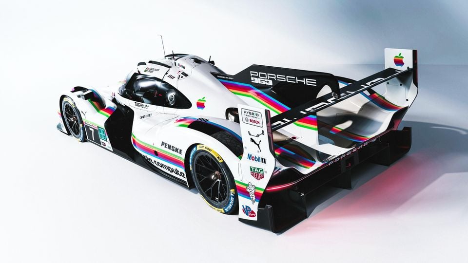 The Porsche 963 LMDh prototype race car in a white livery with rainbow stripes and Apple branding, seen from the rear three-quarter angle in a studio setting