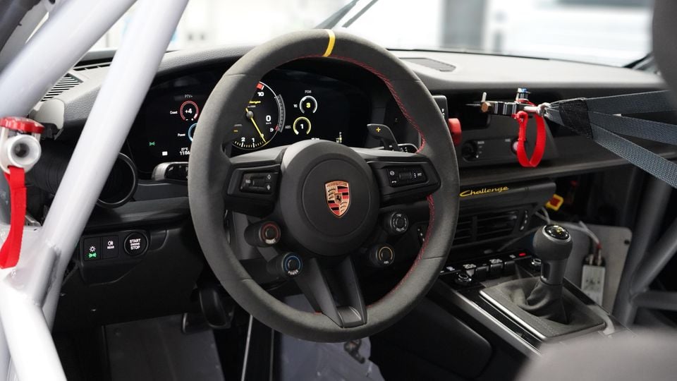 Interior of a Porsche 911 Challenge race car showing the flat-bottom suede steering wheel with Porsche crest, digital instrument cluster, roll cage, and manual gear shifter