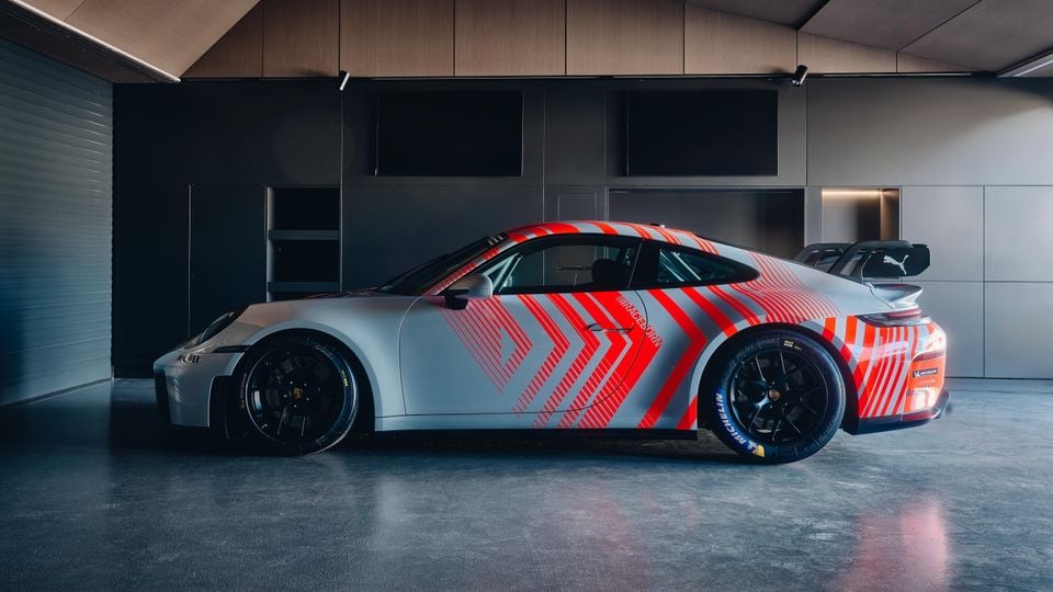 A Porsche 911 GT3 race car with a bold red and grey arrow livery parked inside a modern garage, seen from the side