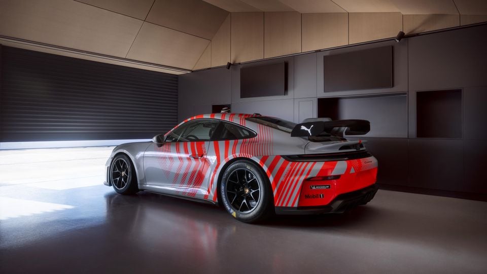 A Porsche 911 GT3 race car with a red and grey livery, featuring a large rear wing, seen from the rear three-quarter angle inside a garage.