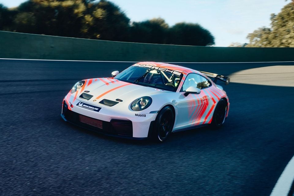 A Porsche 911 GT3 race car with a white and orange livery driving on a circuit track, seen from the front-side