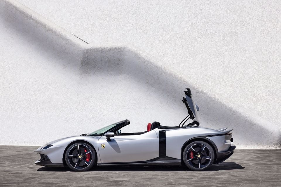 A silver Ferrari supercar seen from the side with the retractable hardtop partially open, parked against a white textured wall