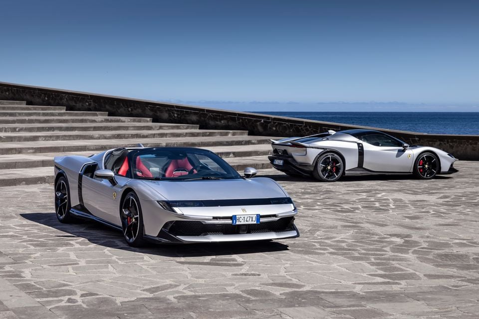 Two silver Ferrari supercars parked on a stone plaza overlooking the sea, with the open-top spider variant in the foreground and a coupe variant behind it