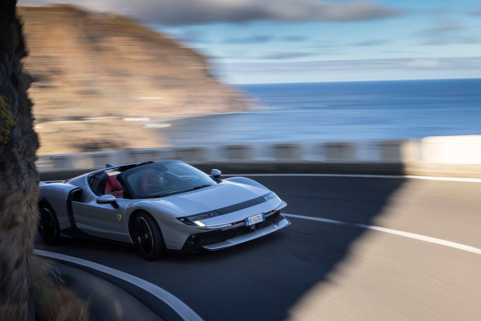 A silver Ferrari open-top supercar driving along a coastal road with cliffs and ocean in the background