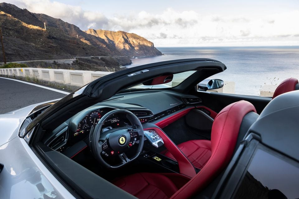 Interior of a white Ferrari convertible showing red leather seats, steering wheel with prancing horse logo, and digital instrument cluster, parked overlooking a coastal mountain road
