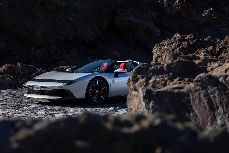 A silver Ferrari open-top supercar parked among rocky terrain, seen from the front-side