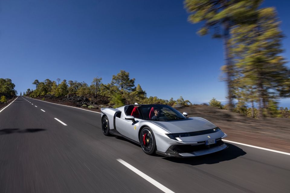 A silver Ferrari open-top supercar being driven along a scenic road lined with trees under a clear blue sky, seen from the front-side
