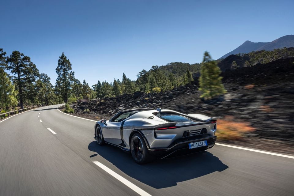 A silver Ferrari supercar seen from the rear, driving along a winding mountain road lined with pine trees under a clear blue sky.