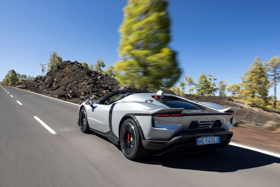 A white Ferrari supercar seen from the rear three-quarter angle, driving along an open road surrounded by trees and rocky terrain under a clear blue sky