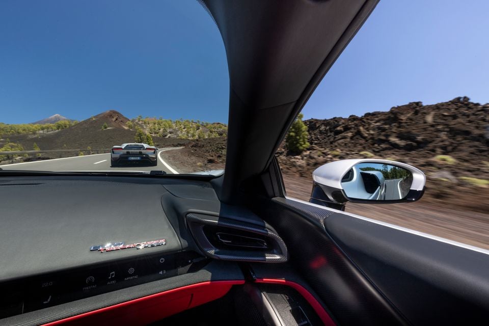 View from inside a supercar's cabin looking through the windscreen at another supercar ahead on a winding mountain road, with volcanic rock and a clear blue sky visible, and the side mirror in the foreground