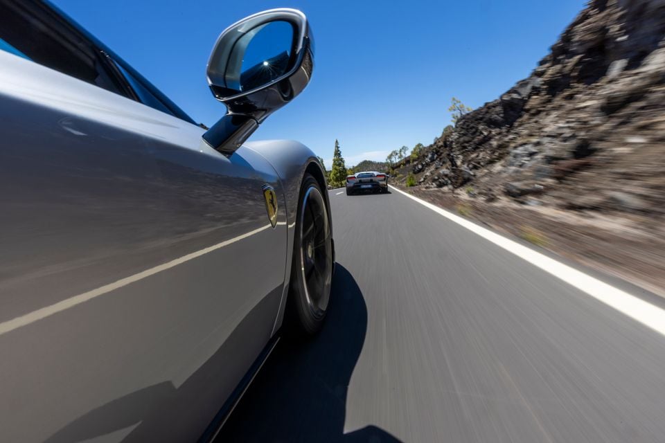 A grey Ferrari driving along a mountain road, with another sports car visible ahead, shot from a low side angle showing the door mirror and front wheel