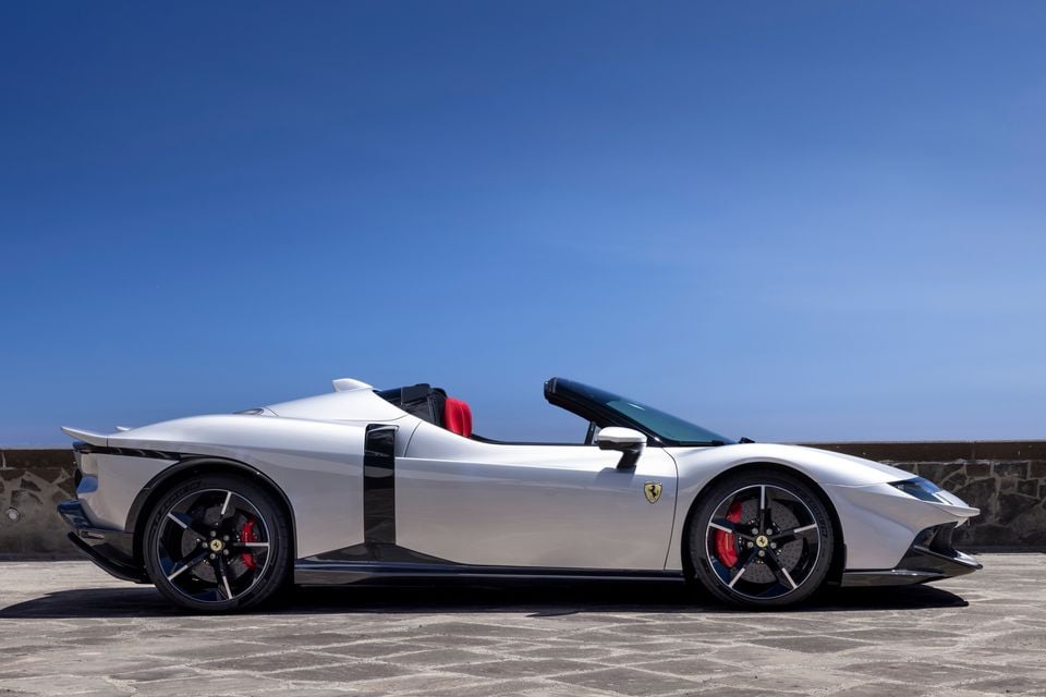A silver Ferrari supercar roadster seen from the side, with an open top, red interior accents, and large black alloy wheels with red brake calipers, parked against a clear blue sky