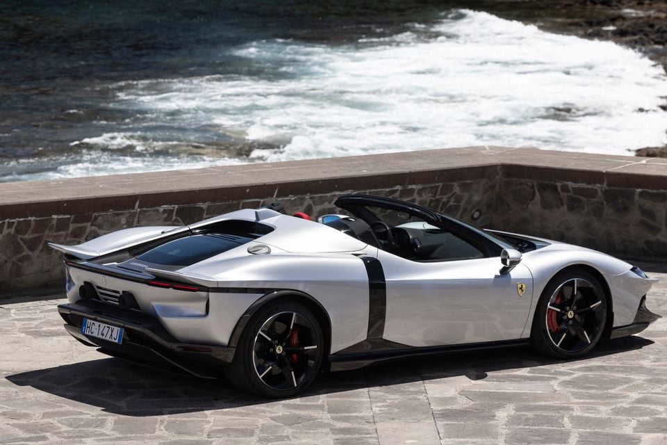 A silver Ferrari spider supercar seen from the rear three-quarter angle, parked on a stone waterfront with the ocean in the background