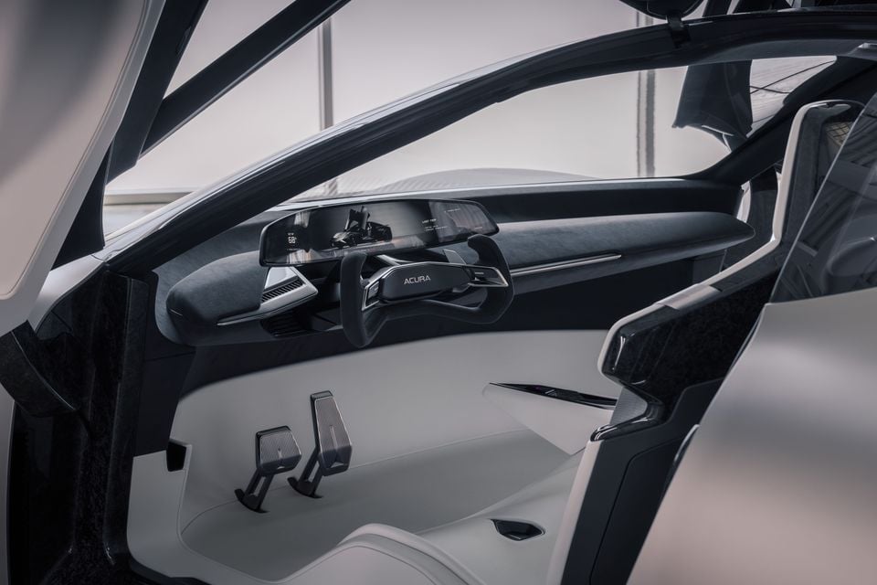 Interior of the Acura Precision EV concept showing a futuristic dashboard, yoke-style steering wheel with Acura branding, wide digital display, and pedals on an open white floor