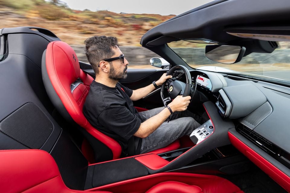 A man driving a Ferrari spider convertible with red leather interior, seen from the side while in motion on an open road