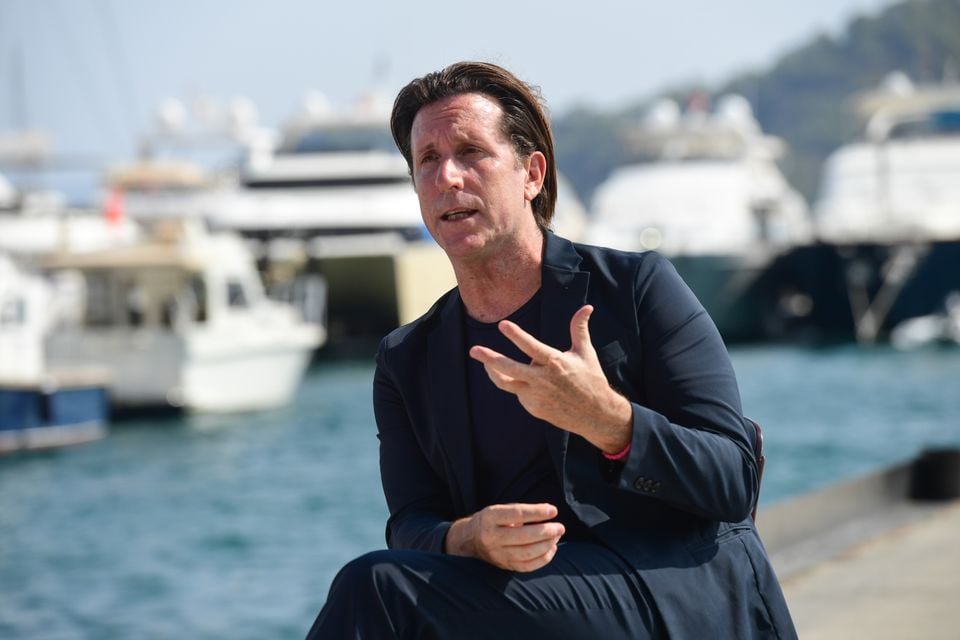 A man in a navy blazer gestures while speaking outdoors at a marina with yachts in the background