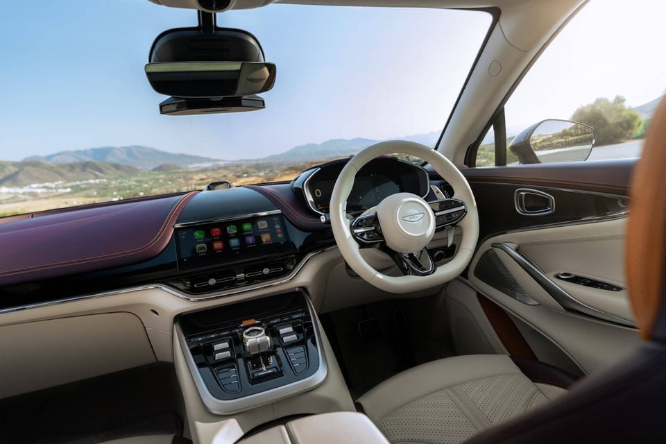The cream and burgundy leather interior of an Aston Martin DBX, showing the steering wheel, dashboard and centre console with mountains visible through the windscreen