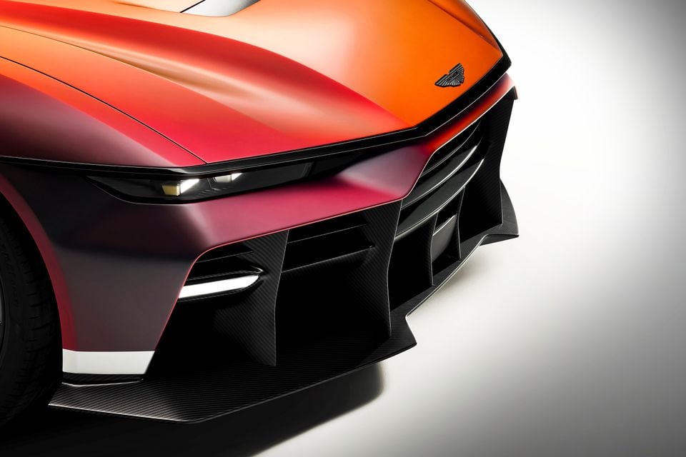 Close-up of the front end of an Aston Martin Valen supercar, showing its aggressive carbon fibre splitter, active headlights, and orange-to-red colour-shifting bodywork in a studio setting.