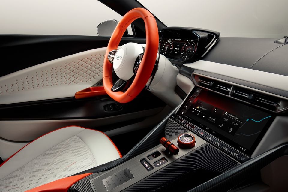 Aston Martin Valen interior showing an orange leather steering wheel, white and orange upholstery, carbon fibre trim, digital instrument cluster, and central touchscreen infotainment system