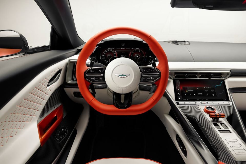 Aston Martin Valen interior showing a red leather steering wheel with Aston Martin badge, digital instrument cluster, central infotainment touchscreen, and white leather trim with orange accents