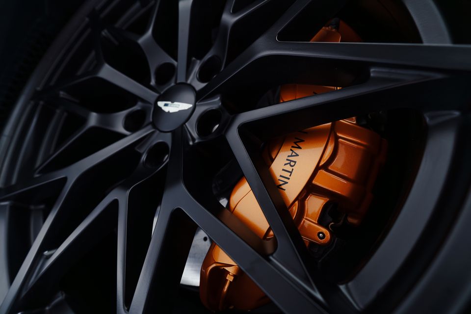 Close-up of an Aston Martin wheel showing a dark multi-spoke alloy rim with an orange brake caliper branded 'Martin'