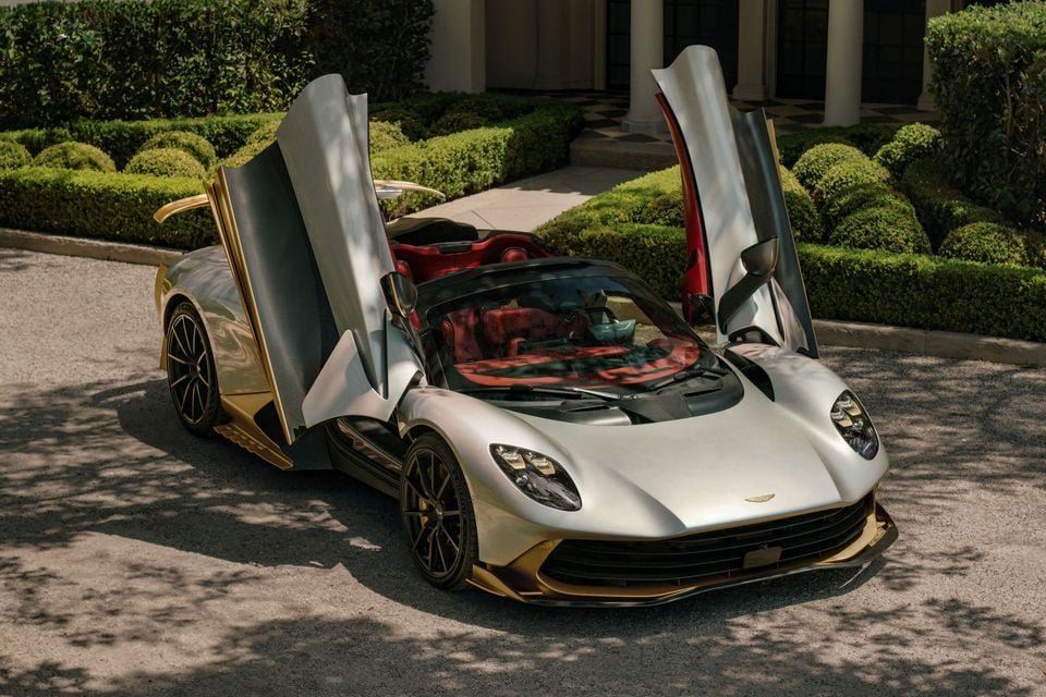 A silver Aston Martin Valhalla Spider with gold accents and its dihedral doors open, parked in front of a manicured garden and mansion